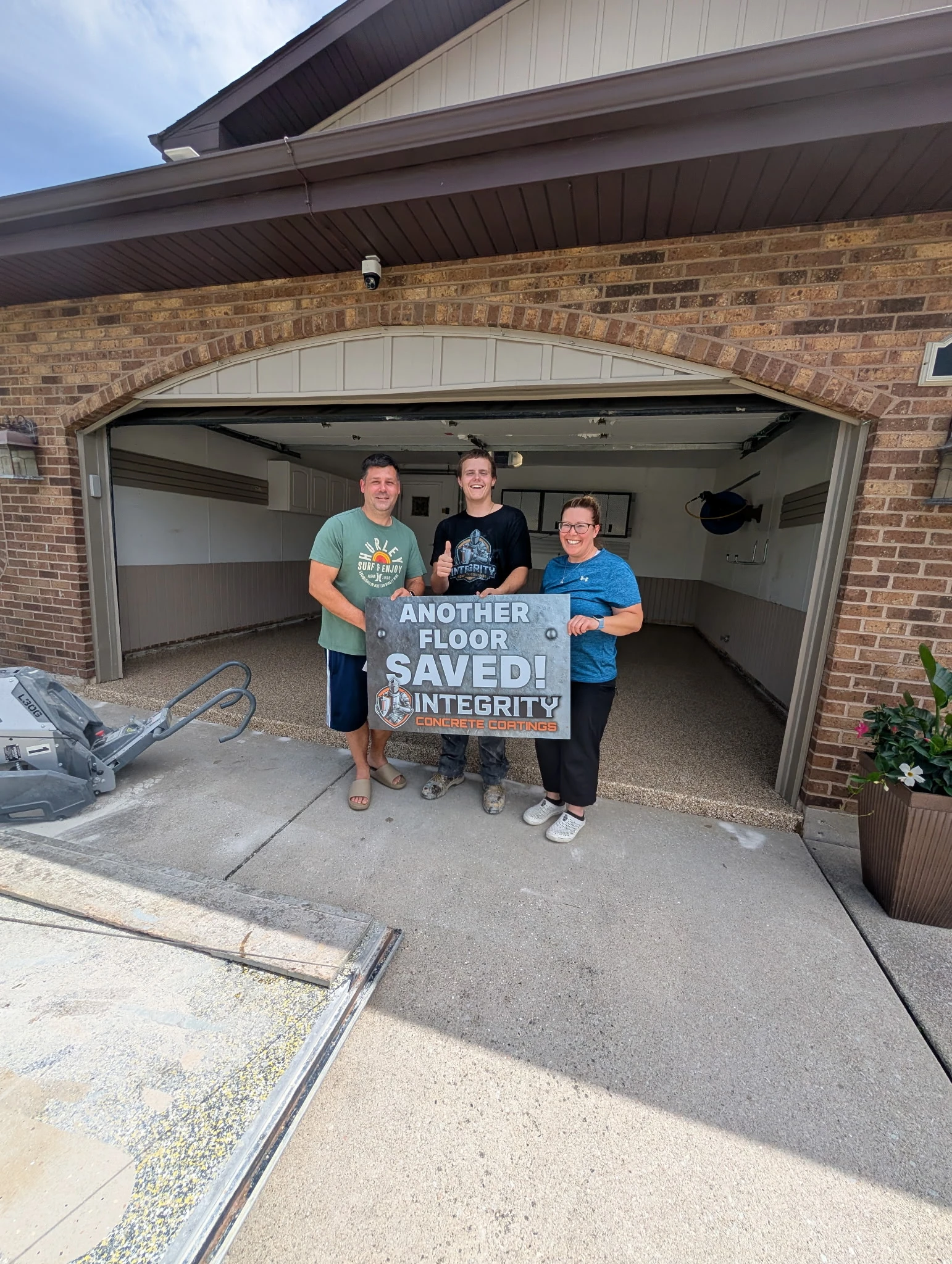 Integrity Concrete Coatings crew member with homeowners holding an Another Floor Saved sign in front of their newly coated garage